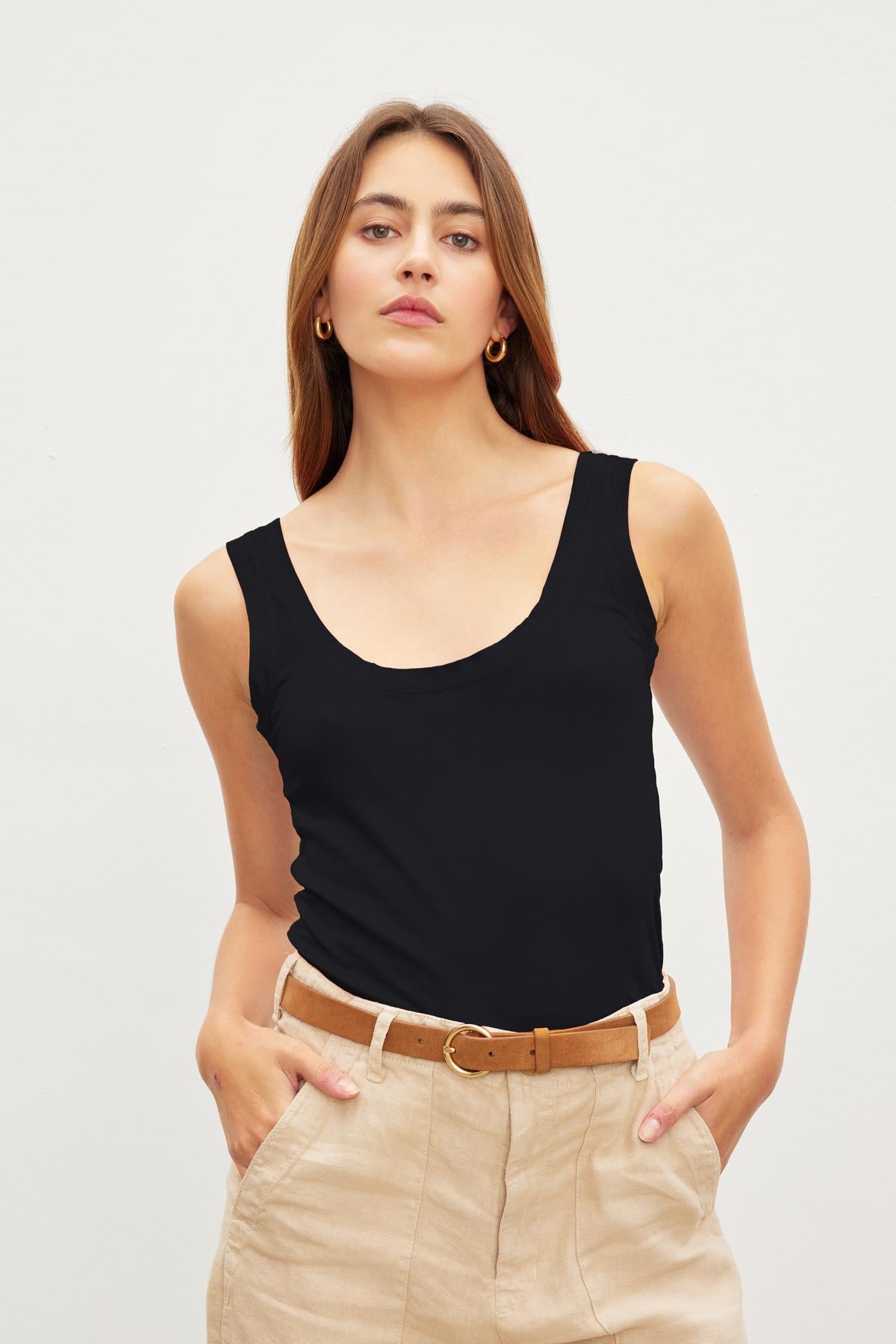 A young woman in a Velvet by Graham & Spencer MOSSY GAUZY WHISPER FITTED TANK and beige trousers with a brown belt, standing against a plain background.-36454076481729