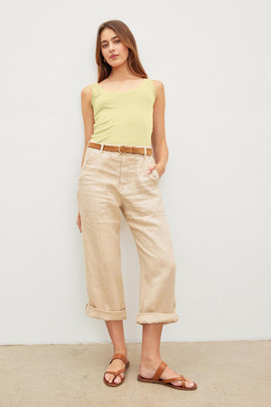 A woman stands posing in a Velvet by Graham & Spencer yellow MOSSY GAUZY WHISPER FITTED TANK, beige cropped pants with a belt, and brown sandals against a plain white background.