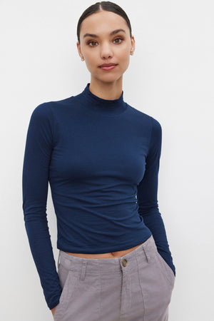 A person wearing the NADENE TEE by Velvet by Graham & Spencer, paired with gray pants, stands with hands in pockets against a plain background.