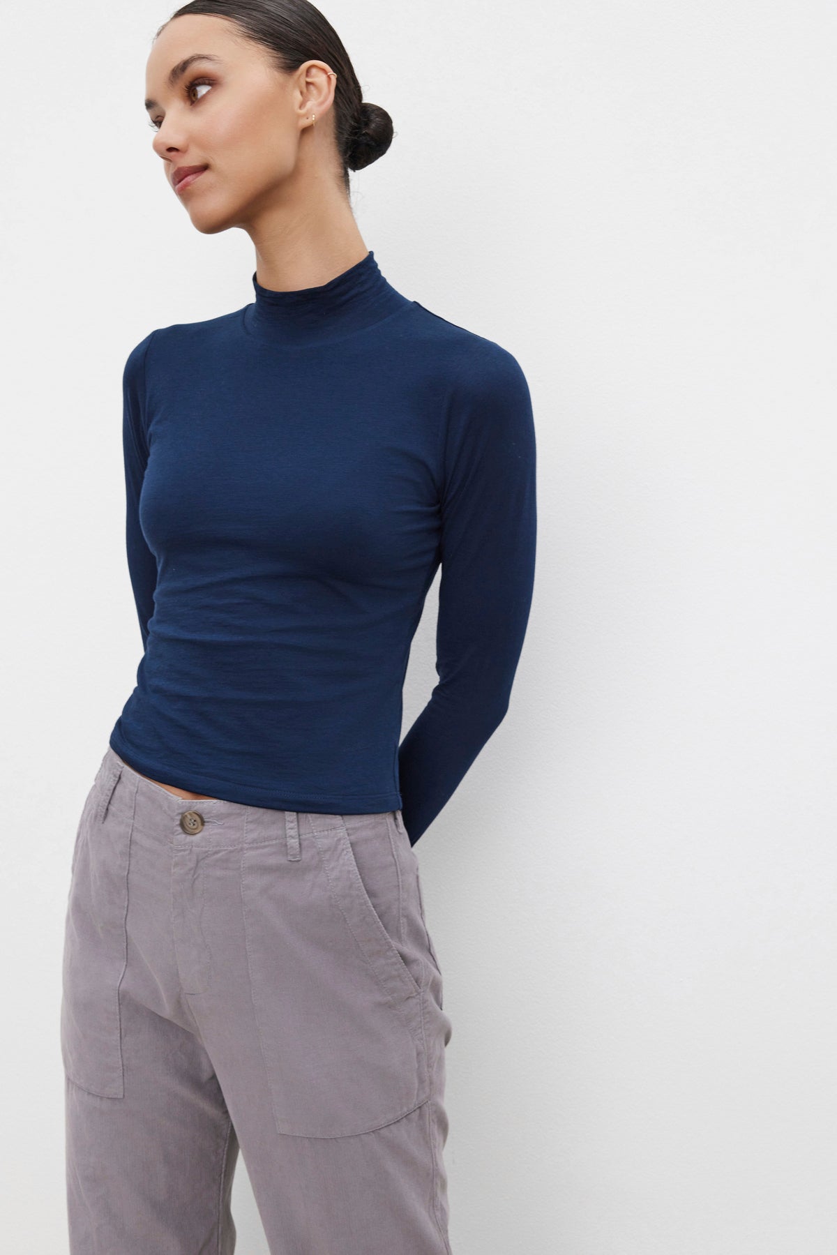   Dressed in a NADENE TEE by Velvet by Graham & Spencer and gray pants, a person poses against a plain white background, gazing to the side with a subtle smile. 