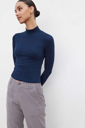 Dressed in a NADENE TEE by Velvet by Graham & Spencer and gray pants, a person poses against a plain white background, gazing to the side with a subtle smile.