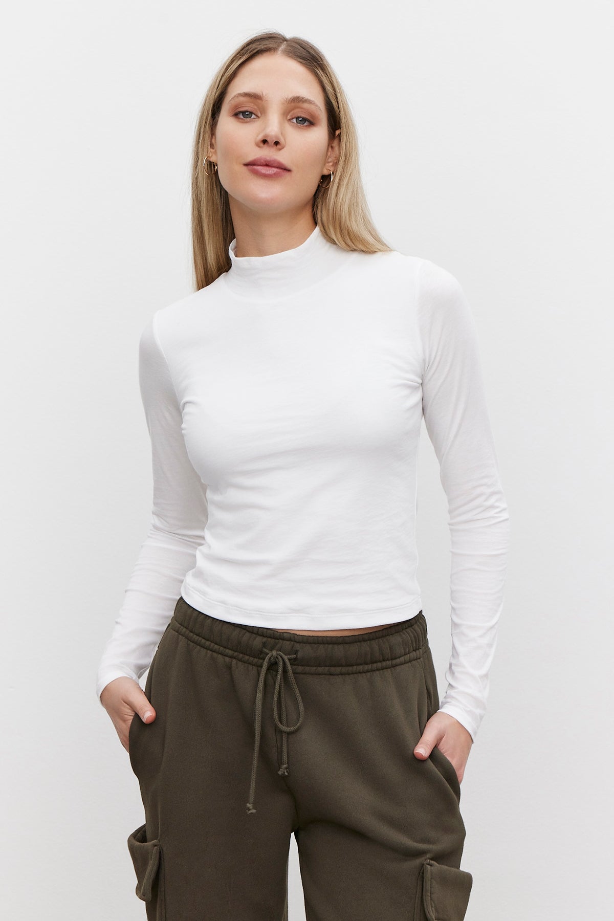   A person wearing the NADENE TEE by Velvet by Graham & Spencer, a white lightweight long-sleeve top, paired with olive green pants stands against a plain background with hands casually in pockets. 