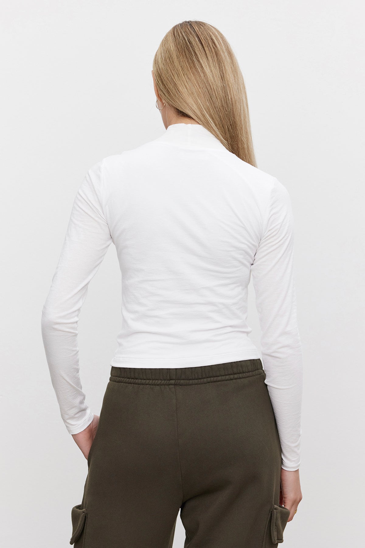   A person with long blonde hair is seen from the back wearing the NADENE TEE, a white mock neck, long-sleeve shirt from Velvet by Graham & Spencer, paired with green pants in front of a plain background. 