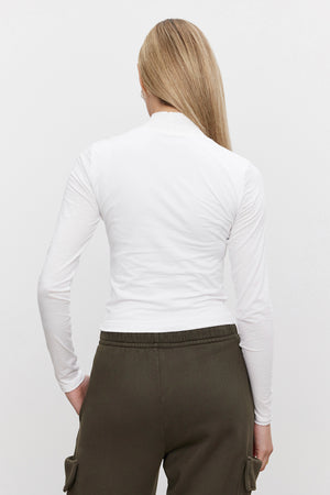 A person with long blonde hair is seen from the back wearing the NADENE TEE, a white mock neck, long-sleeve shirt from Velvet by Graham & Spencer, paired with green pants in front of a plain background.