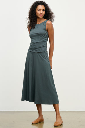 A woman wears the PIA DRESS by Velvet by Graham & Spencer— a sleeveless, dark gray midi with round neckline, fitted bodice, and flared skirt—paired with tan flats against a plain light background.