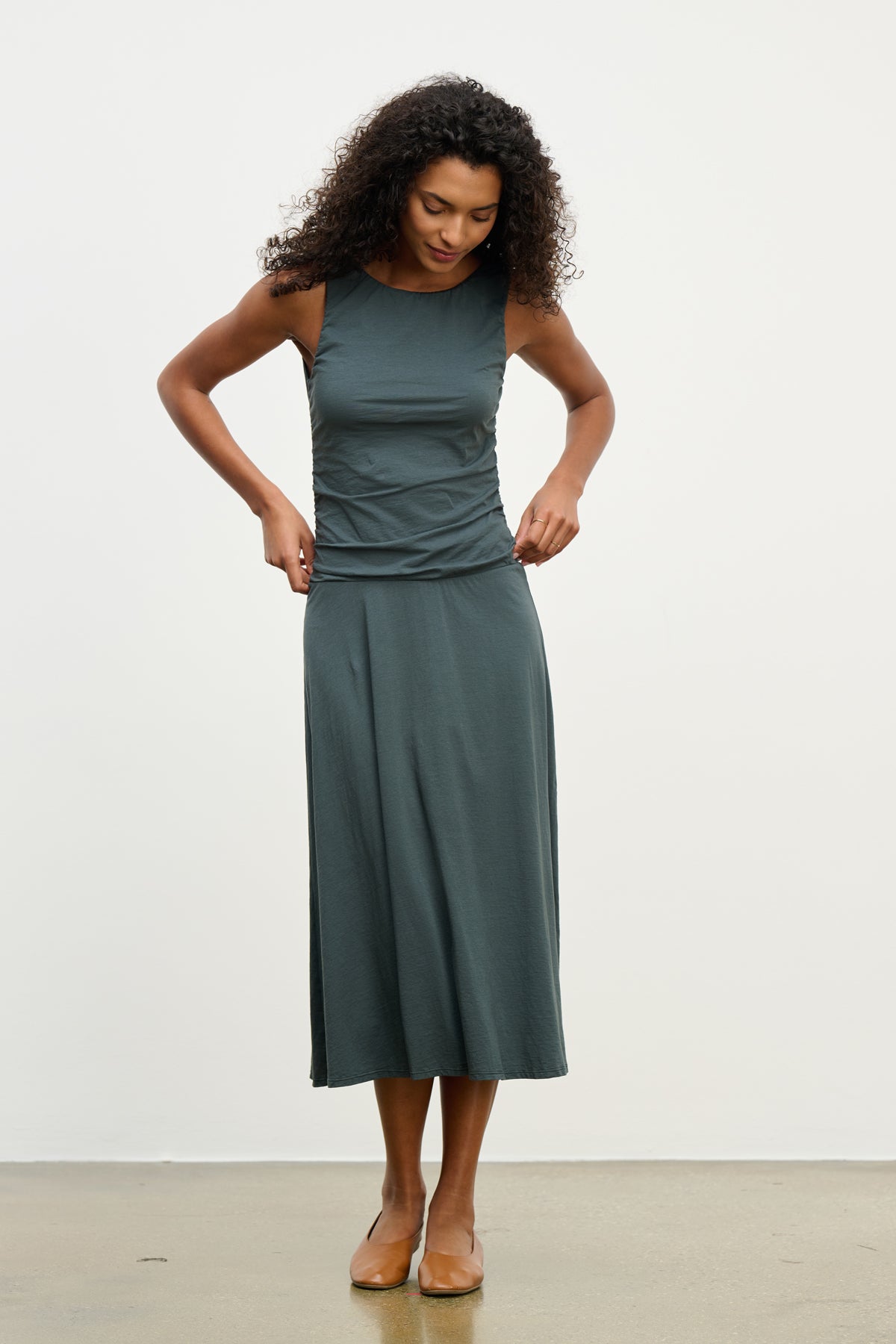   A woman wearing the PIA DRESS by Velvet by Graham & Spencer—a sleeveless dark green midi dress with a round neckline—stands indoors on a light-colored floor, looking down as she adjusts her outfit. 