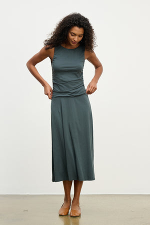A woman wearing the PIA DRESS by Velvet by Graham & Spencer—a sleeveless dark green midi dress with a round neckline—stands indoors on a light-colored floor, looking down as she adjusts her outfit.
