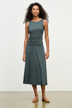 A woman with curly hair stands against a plain background, wearing the PIA DRESS by Velvet by Graham & Spencer—a sleeveless, dark green midi dress with a round neckline—and brown flat shoes.