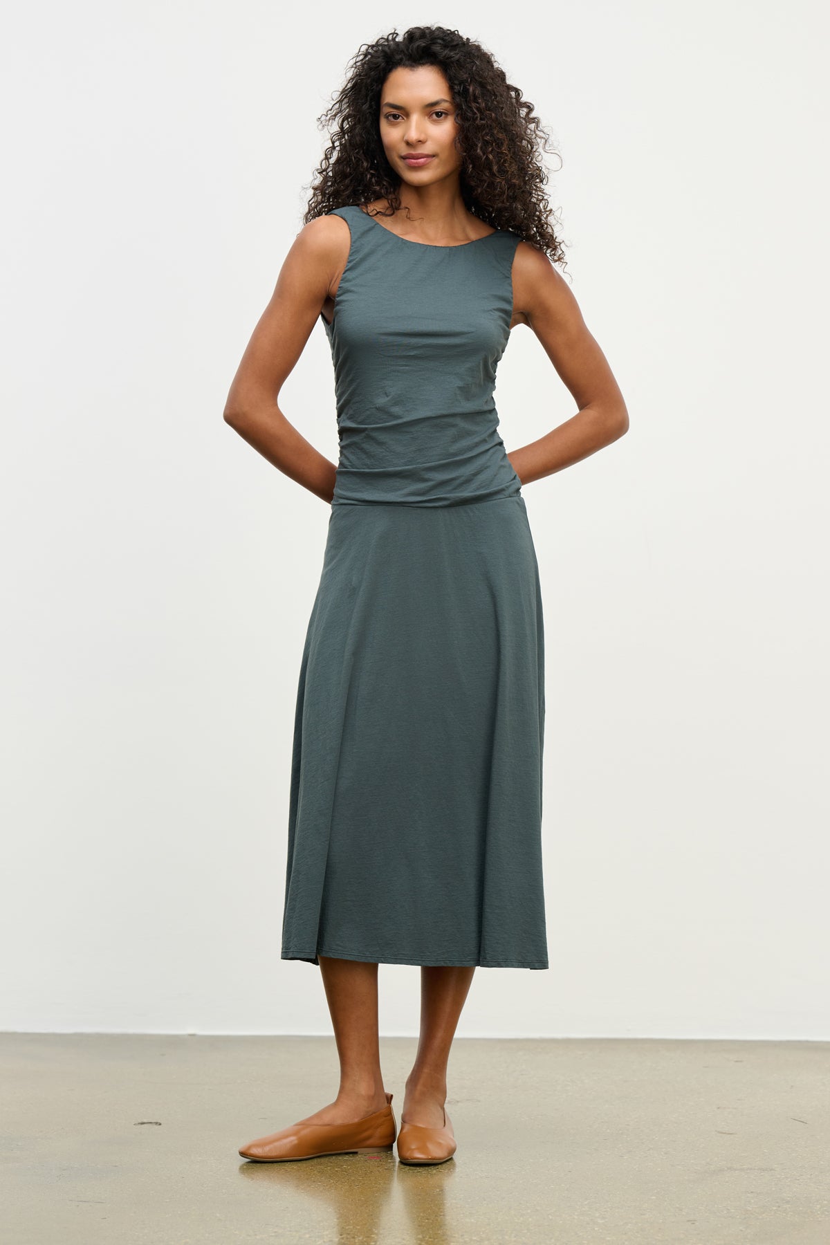 A woman stands against a plain background wearing the Velvet by Graham & Spencer PIA DRESS, a sleeveless dark green lined midi dress with a round neckline, paired with brown flat shoes.-39538942181569