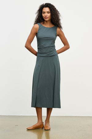 A woman stands against a plain background wearing the Velvet by Graham & Spencer PIA DRESS, a sleeveless dark green lined midi dress with a round neckline, paired with brown flat shoes.