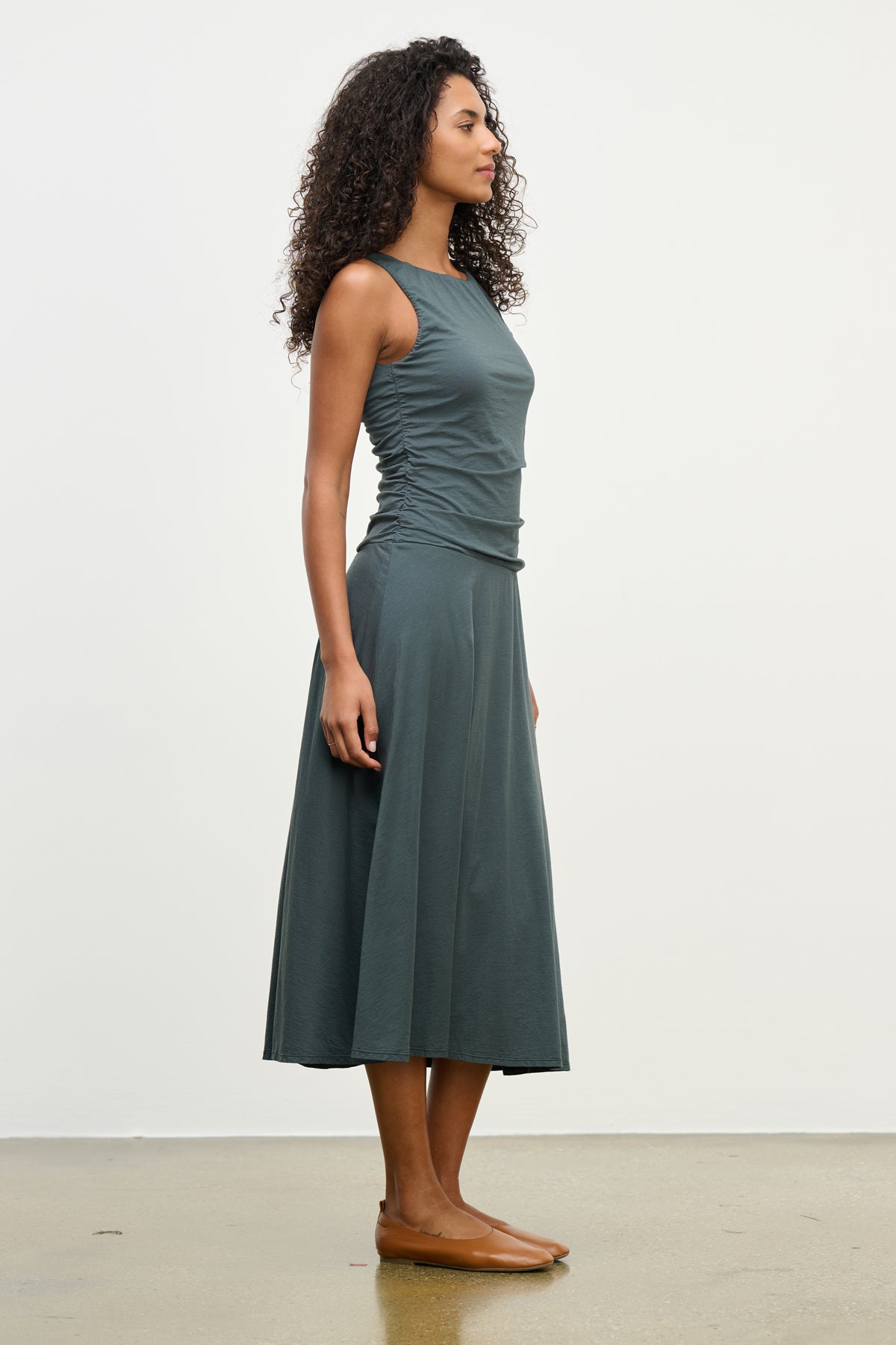 A woman stands in profile against a white background, wearing Velvet by Graham & Spencer's PIA DRESS—a sleeveless, lined, ankle-length dark gray dress with a round neckline—paired with brown flats.-39538942148801