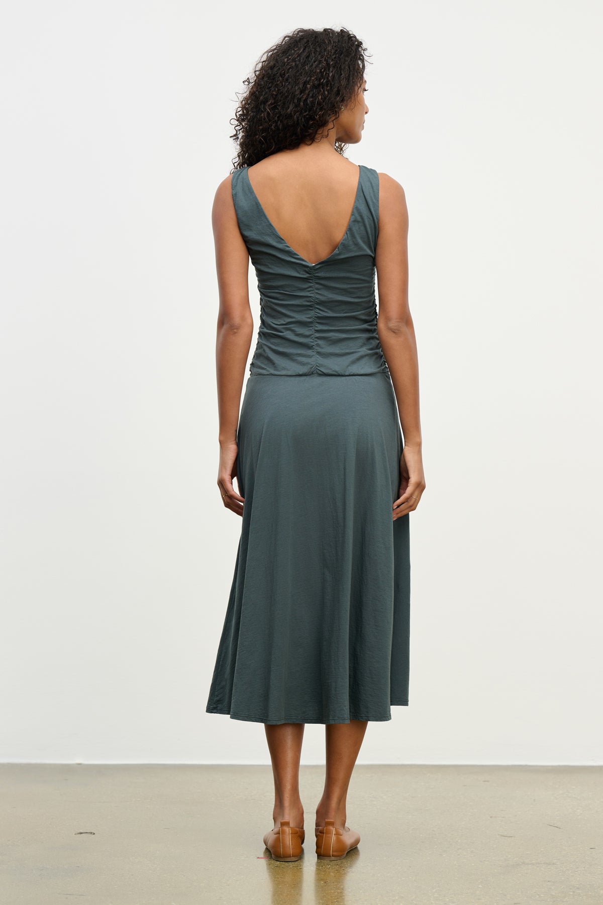 A woman faces away wearing the PIA DRESS by Velvet by Graham & Spencer—a sleeveless, dark teal midi dress with a V-back and ruched details. The lined dress is styled with brown sandals against a simple background.-39538942116033
