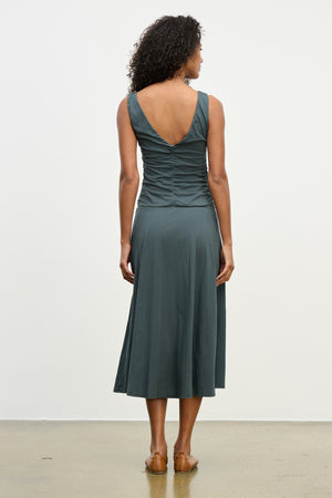 A woman faces away wearing the PIA DRESS by Velvet by Graham & Spencer—a sleeveless, dark teal midi dress with a V-back and ruched details. The lined dress is styled with brown sandals against a simple background.