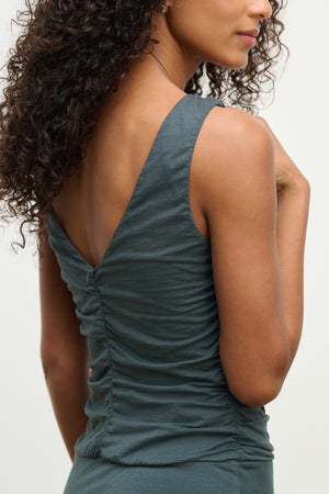 A person with curly hair is shown from the back, wearing the PIA DRESS by Velvet by Graham & Spencer—a sleeveless, ruched, dark teal lined dress featuring a V-shaped neckline.