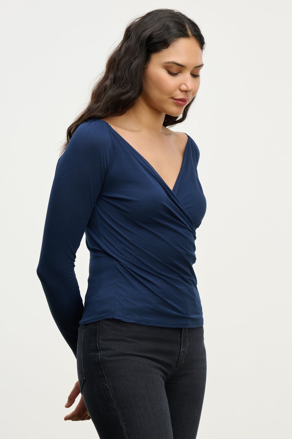 Woman with long dark hair wearing the RAK TEE by Velvet by Graham & Spencer, a navy blue wrap-front top with a feminine silhouette, paired with black jeans, standing with her hands behind her back and looking down.-39815250936001