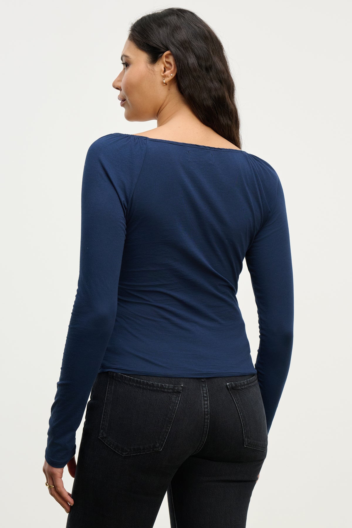 A woman with long dark hair, wearing the Velvet by Graham & Spencer RAK TEE and black jeans, stands with her back to the camera against a plain background, showcasing a feminine silhouette.-39815250903233