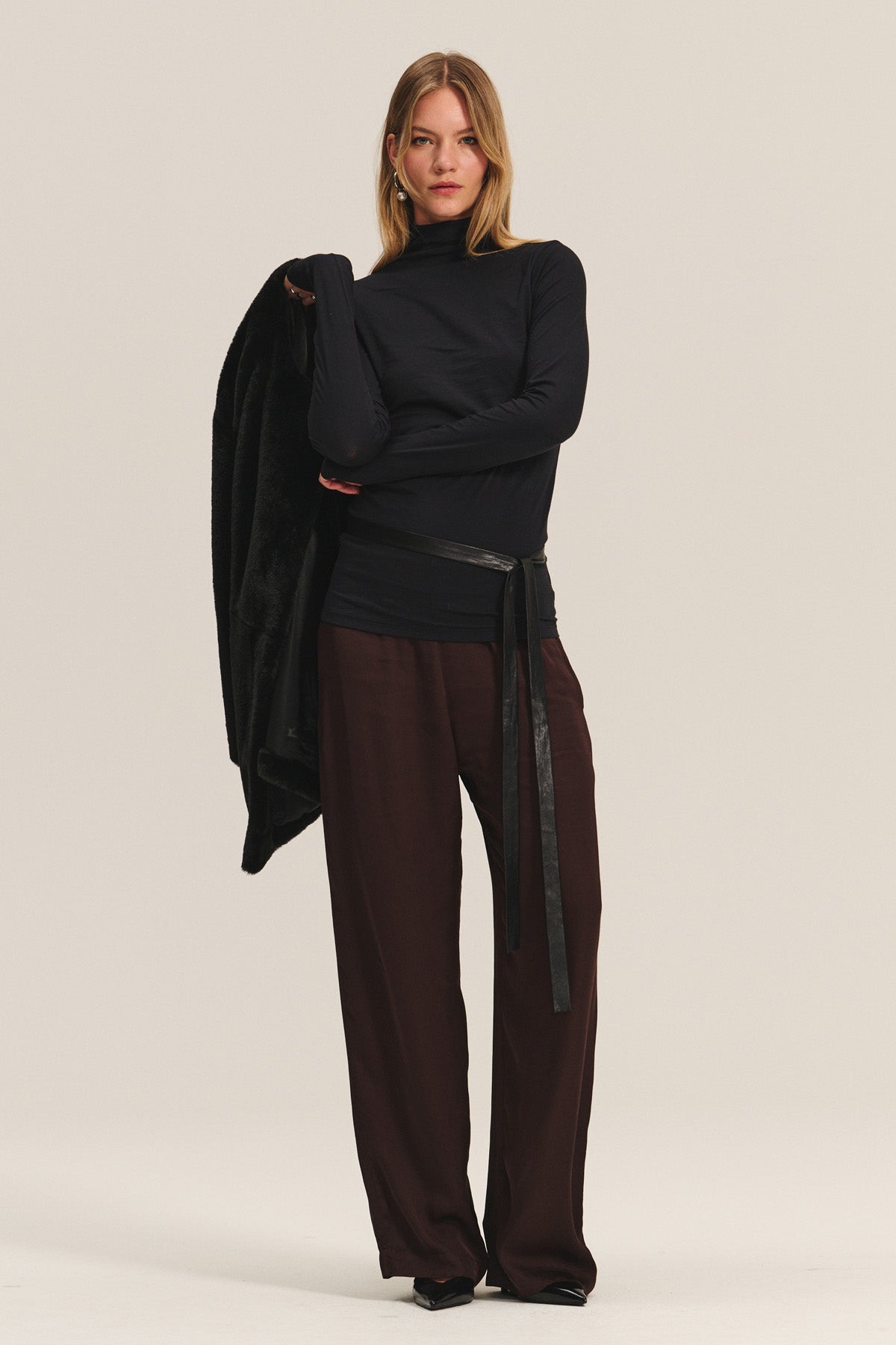   A person stands facing forward, wearing a black turtleneck, black belt, and the Velvet by Graham & Spencer LARKIN PANT in dark brown satin with a flattering wide-leg fit, holding a black jacket over one arm against a plain background. 
