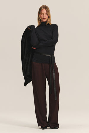 A person stands facing forward, wearing a black turtleneck, black belt, and the Velvet by Graham & Spencer LARKIN PANT in dark brown satin with a flattering wide-leg fit, holding a black jacket over one arm against a plain background.
