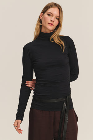 A woman in the TALISIA TEE by Velvet by Graham & Spencer—a black mock neck long-sleeve top—pairs it with brown pants and long earrings, standing against a plain light background for a timeless look.