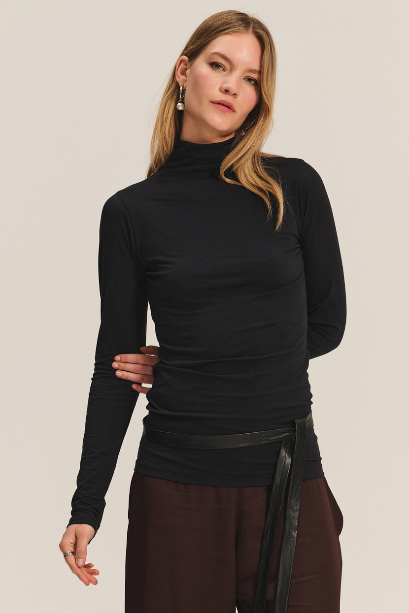 A woman in the TALISIA TEE by Velvet by Graham & Spencer—a black mock neck long-sleeve top—pairs it with brown pants and long earrings, standing against a plain light background for a timeless look.
