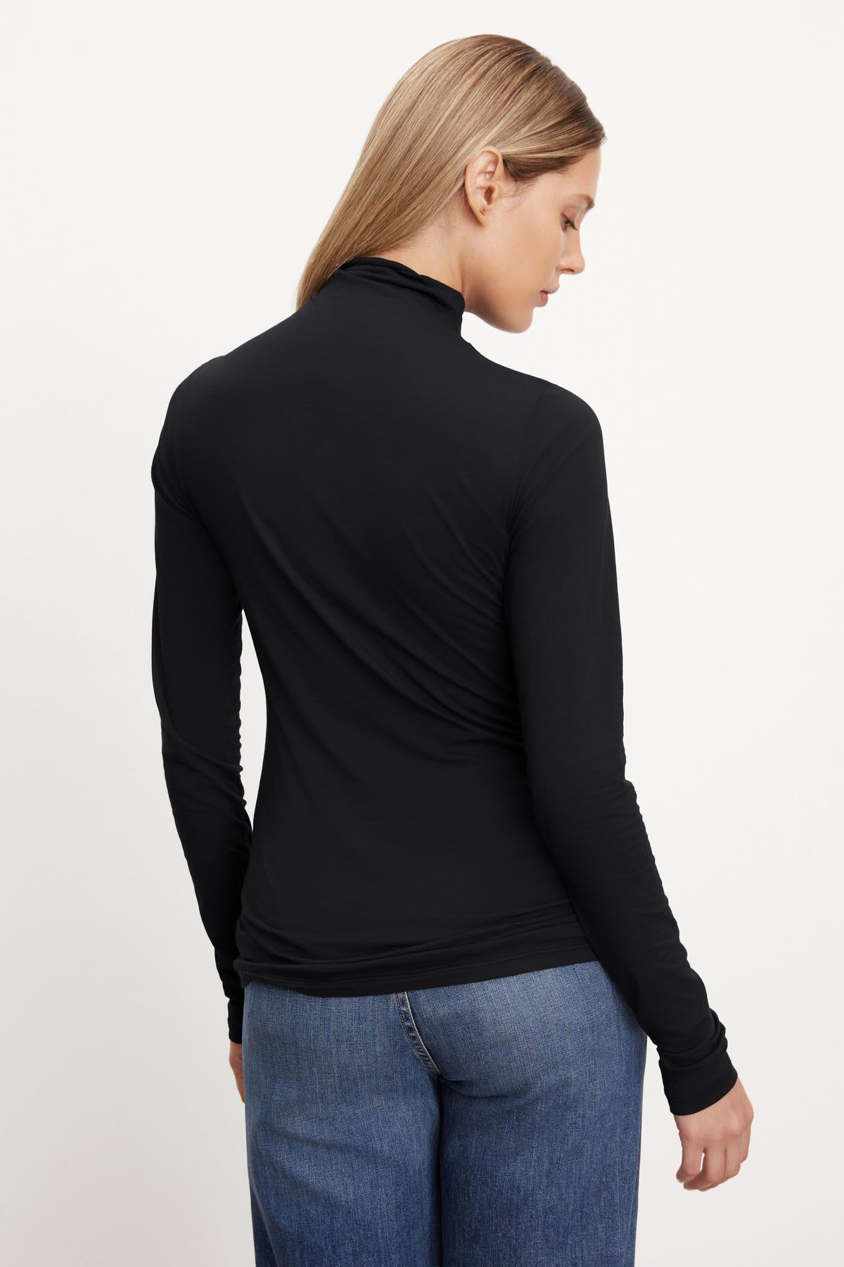   A person with long blonde hair stands facing away, showcasing the TALISIA TEE by Velvet by Graham & Spencer—a versatile black long-sleeve turtleneck that epitomizes timeless fashion. Paired with blue jeans against a plain backdrop, this look effortlessly combines elegance and simplicity. 