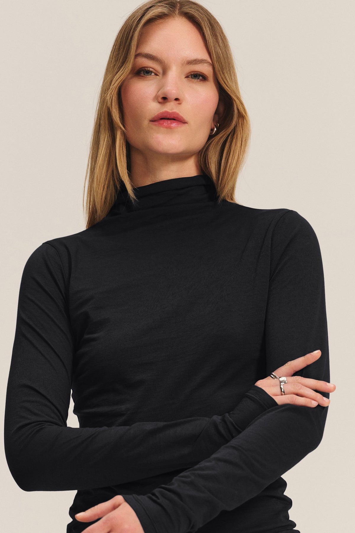 A woman with straight blonde hair stands against a neutral background, arms folded, wearing the TALISIA TEE by Velvet by Graham & Spencer—a black mock neck long-sleeve top that's a timeless wardrobe staple.-40182502162625