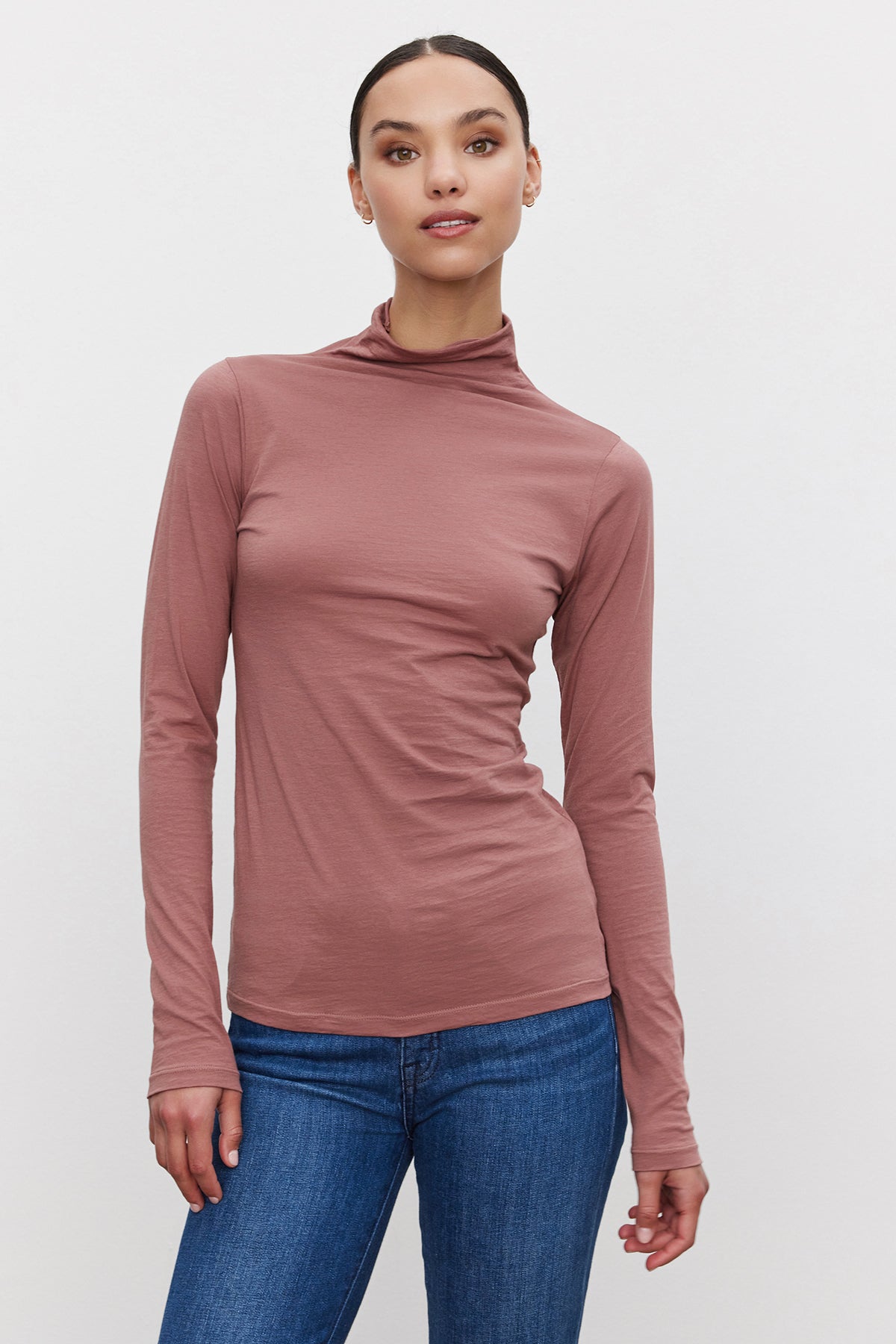   A person wearing the TALISIA TEE, a rose-colored, long-sleeved mock neck shirt by Velvet by Graham & Spencer, paired with blue jeans is standing against a plain white background, showcasing their fashionable and versatile wardrobe. 