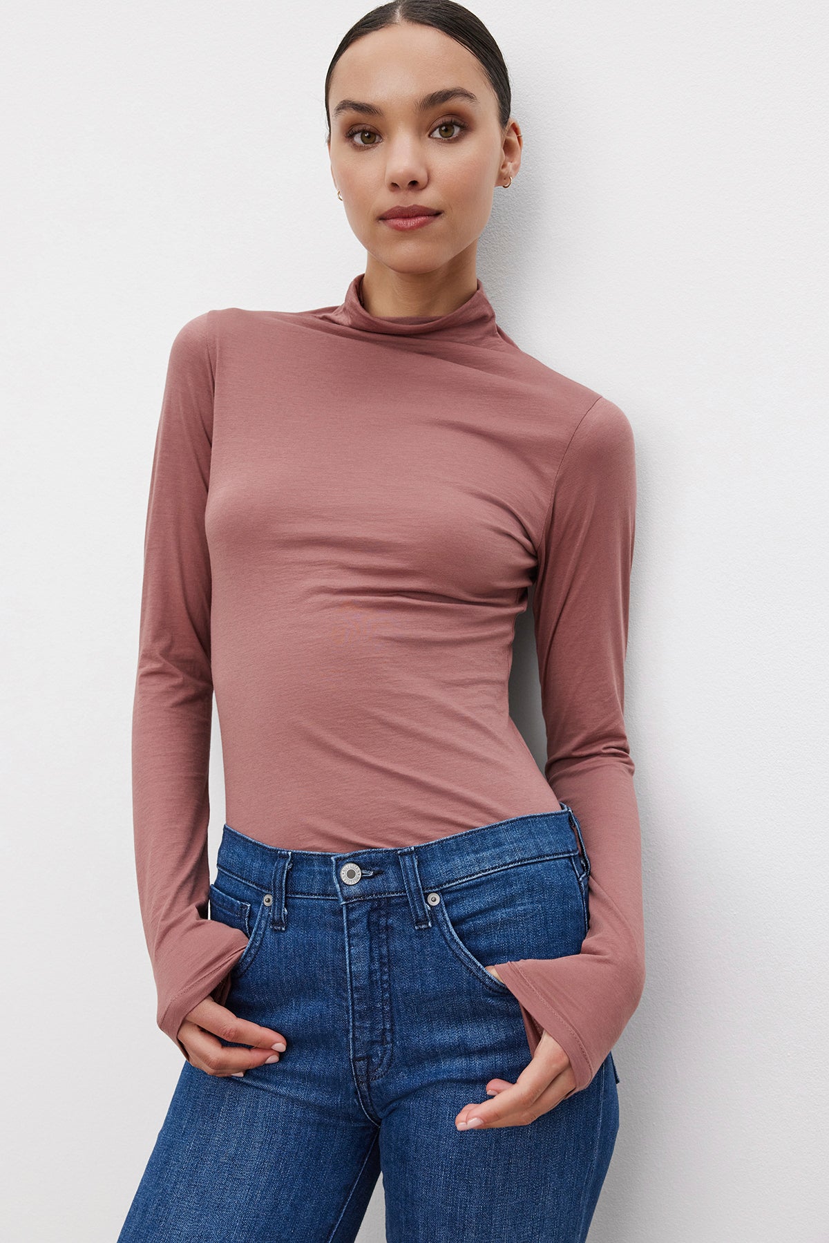 A woman stands against a white background wearing the stylish, long-sleeved, mock-neck TALISIA TEE in mauve by Velvet by Graham & Spencer paired with blue jeans. With her hands in her pockets, she looks calmly at the camera, showcasing a versatile wardrobe choice for any season.-37855992217793