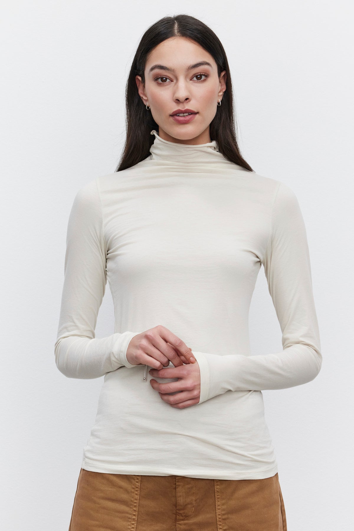   A person wearing a versatile wardrobe staple: the long-sleeved, cream-colored TALISIA TEE by Velvet by Graham & Spencer and brown pants, stands against a plain white background. 