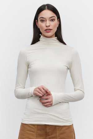 A person wearing a versatile wardrobe staple: the long-sleeved, cream-colored TALISIA TEE by Velvet by Graham & Spencer and brown pants, stands against a plain white background.