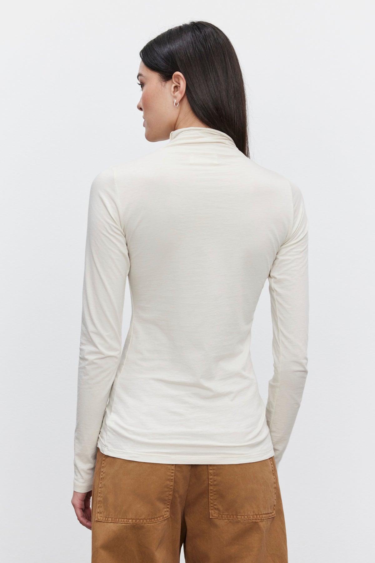   Rear view of a person with long dark hair wearing the Velvet by Graham & Spencer TALISIA TEE, a cream-colored mock neck long-sleeve turtleneck paired with brown pants, standing against a plain white background. A versatile wardrobe staple for any fashion enthusiast. 