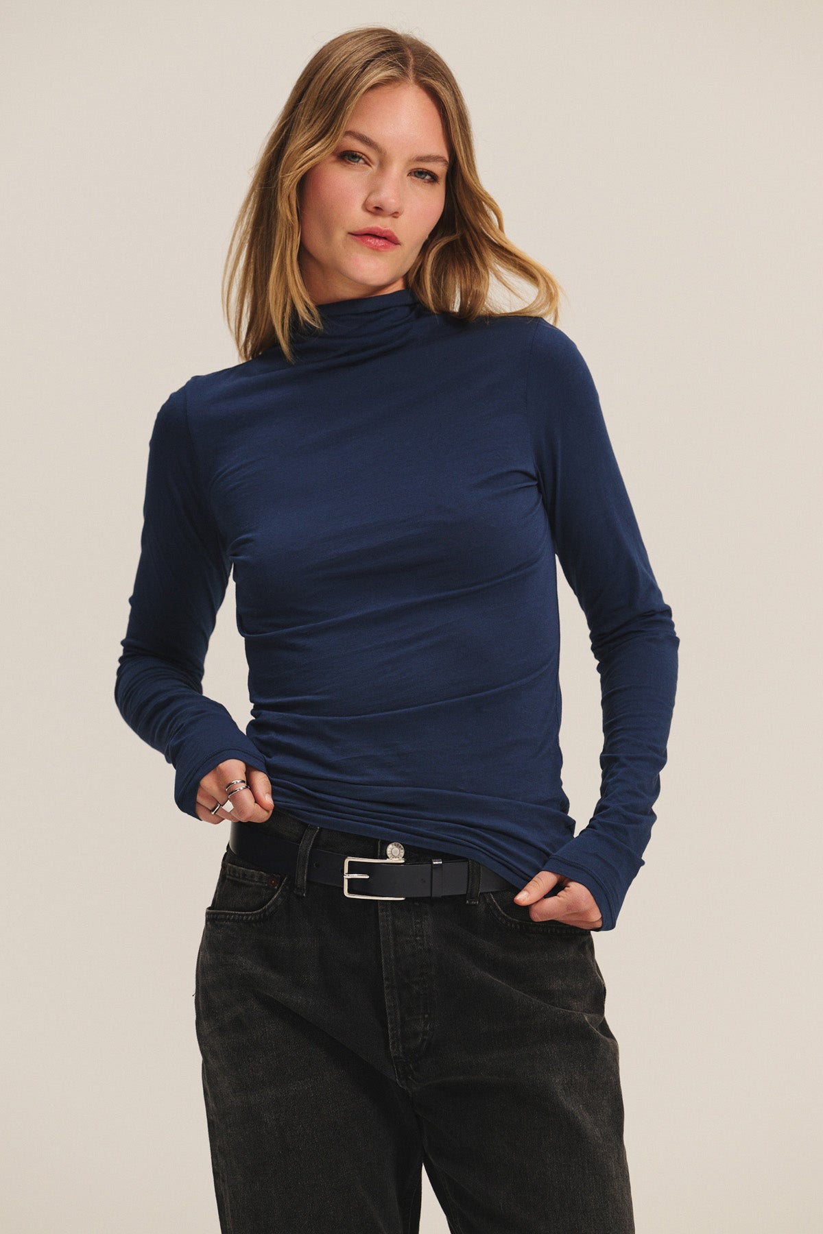 A woman wears the Velvet by Graham & Spencer TALISIA TEE, a fitted navy mock neck long-sleeve top—styled with black jeans and a black belt against a plain background; this piece is a true wardrobe staple.-40252512370881