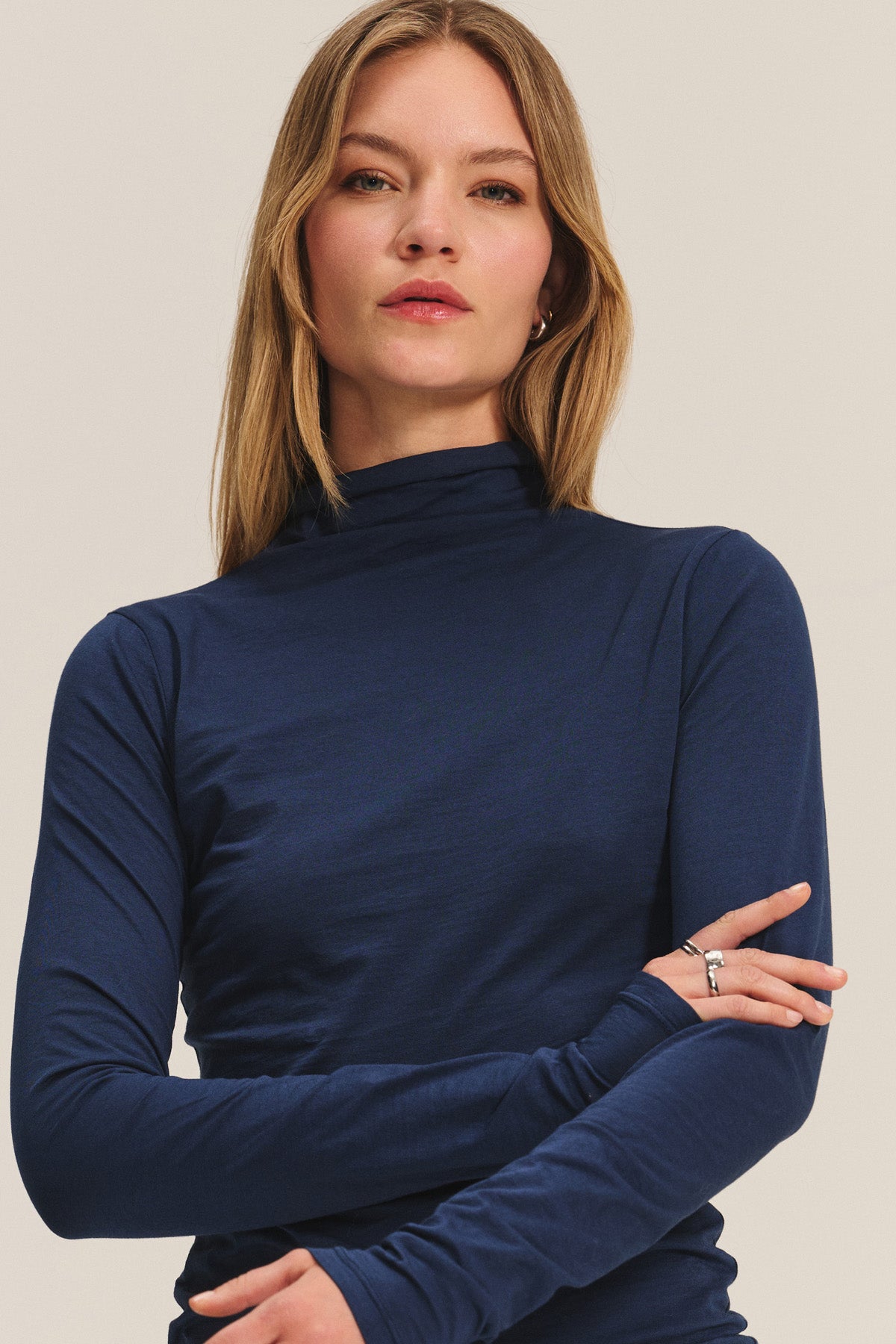 A woman with straight, light brown hair wears the TALISIA TEE by Velvet by Graham & Spencer—a fitted, long-sleeve navy blue mock neck top—paired with minimal jewelry, standing against a neutral background.-40252512305345