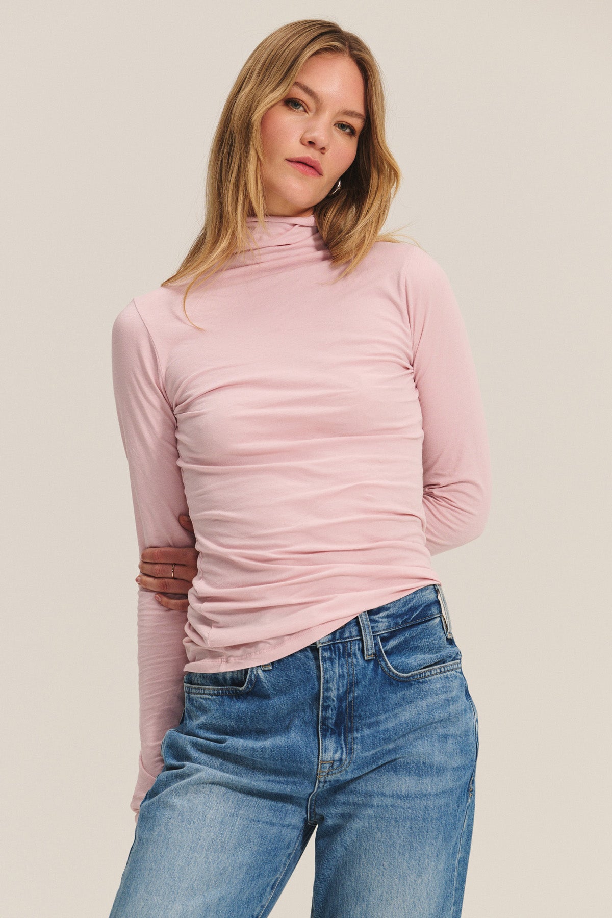 A person wears the TALISIA TEE by Velvet by Graham & Spencer—a light pink mock neck long-sleeve top—paired with blue jeans, standing against a plain light background to showcase this timeless wardrobe staple.-40248255054017