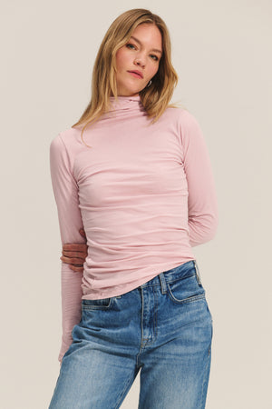 A person wears the TALISIA TEE by Velvet by Graham & Spencer—a light pink mock neck long-sleeve top—paired with blue jeans, standing against a plain light background to showcase this timeless wardrobe staple.