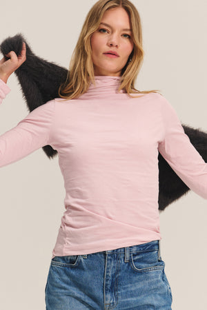A woman faces forward, wearing the TALISIA TEE by Velvet by Graham & Spencer in light pink with blue jeans, holding a dark jacket over her shoulders against a plain background.