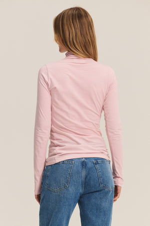 A person with light brown hair is seen from the back wearing Velvet by Graham & Spencer’s TALISIA TEE in light pink with blue jeans, creating a simple wardrobe staple look against a plain background.