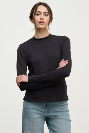 A person stands against a plain background in the TRISH TEE by Velvet by Graham & Spencer and light blue jeans, arms crossed and expression neutral—an effortlessly stylish look for everyday wear.
