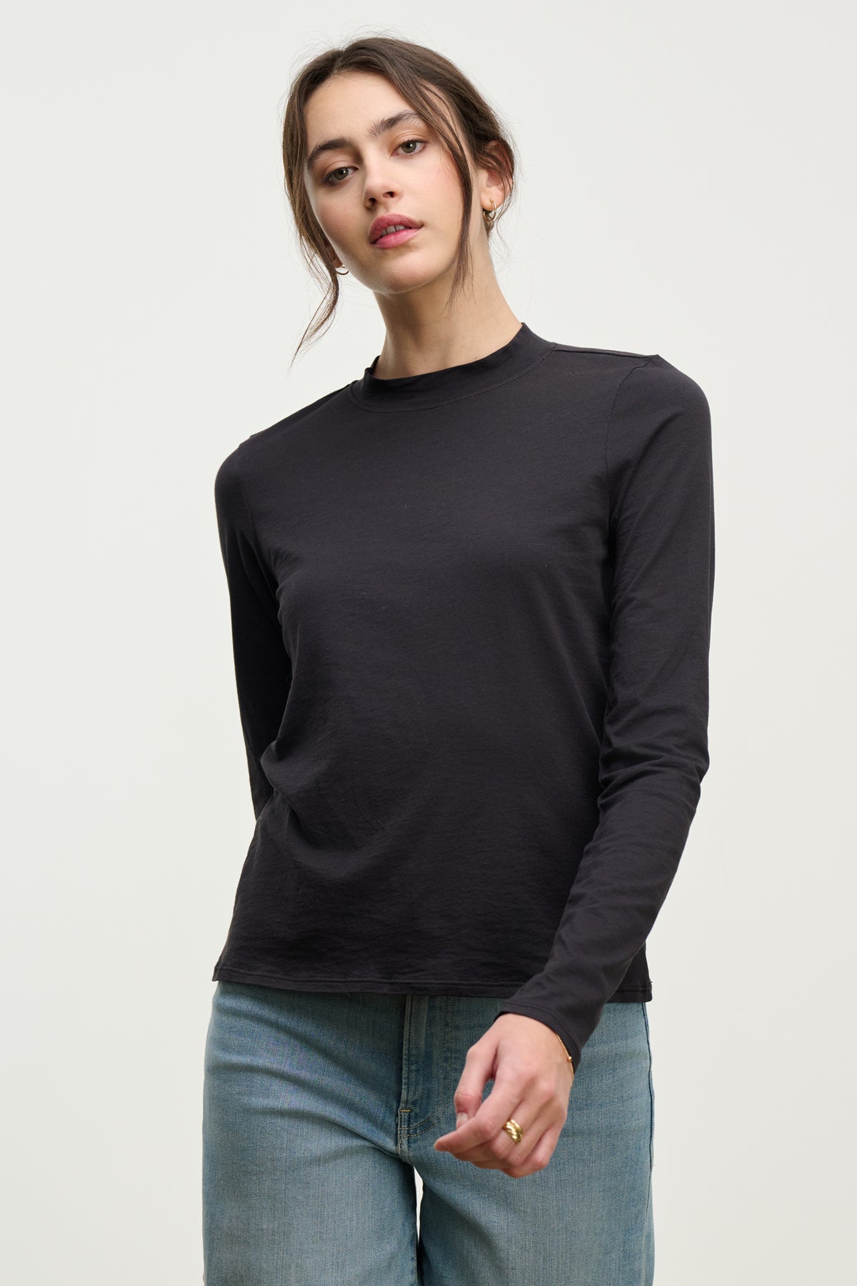   A woman wears the TRISH TEE by Velvet by Graham & Spencer in black with light blue jeans, standing against a plain light background and showcasing simple everyday style. 