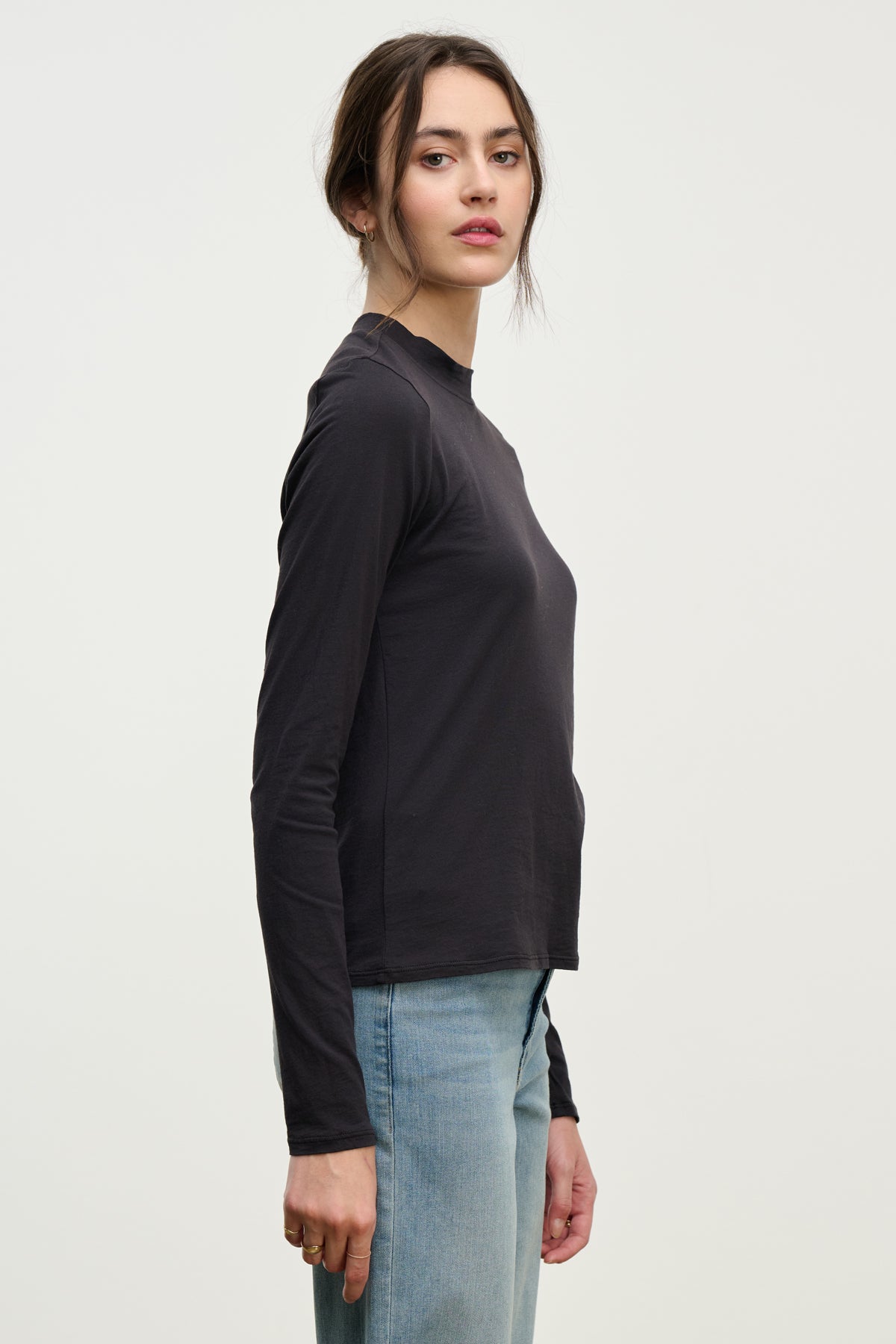   A woman stands sideways in the TRISH TEE by Velvet by Graham & Spencer—a long-sleeve black crewneck top—paired with light blue jeans, her hair tied back and a neutral expression, set against a plain white background for an effortless everyday look. 