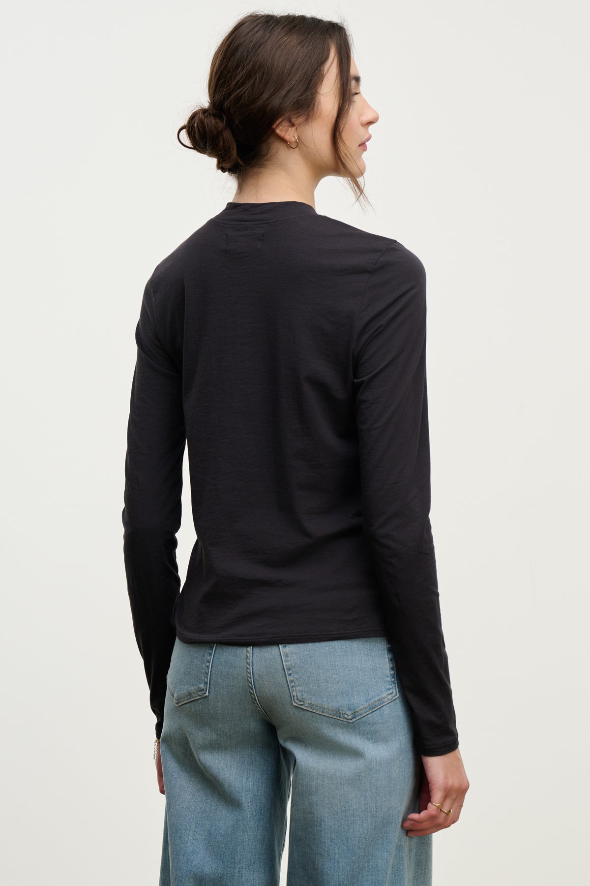   A woman with brown hair in a low bun wears the Velvet by Graham & Spencer TRISH TEE—a black, long-sleeve lightweight crewneck—paired with light blue jeans, standing with her back to the camera against a plain white background. 