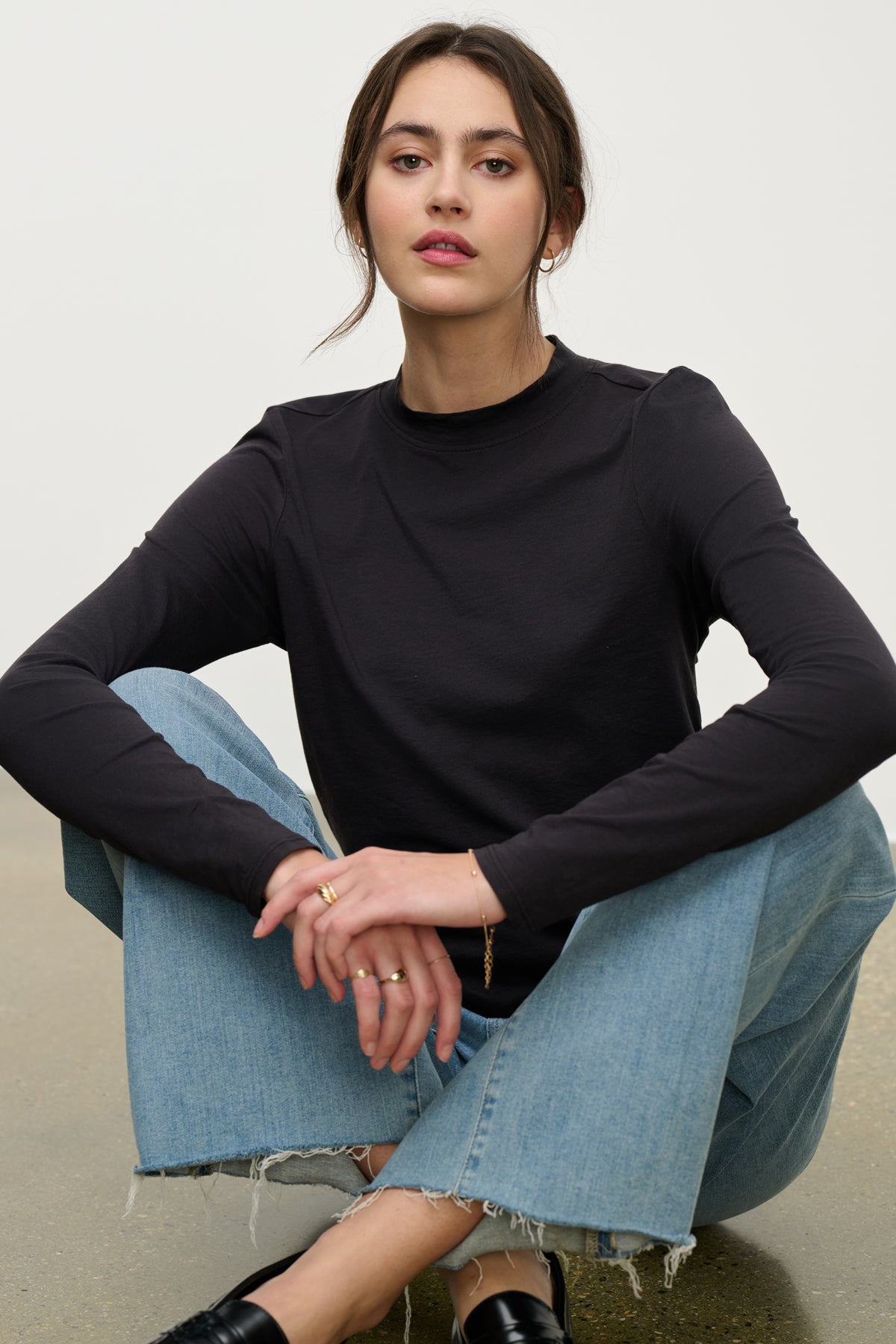 A woman wears the TRISH TEE by Velvet by Graham & Spencer, a black crew neck long-sleeve shirt paired with light blue jeans, sitting cross-legged on the floor and looking at the camera—perfect for everyday style.