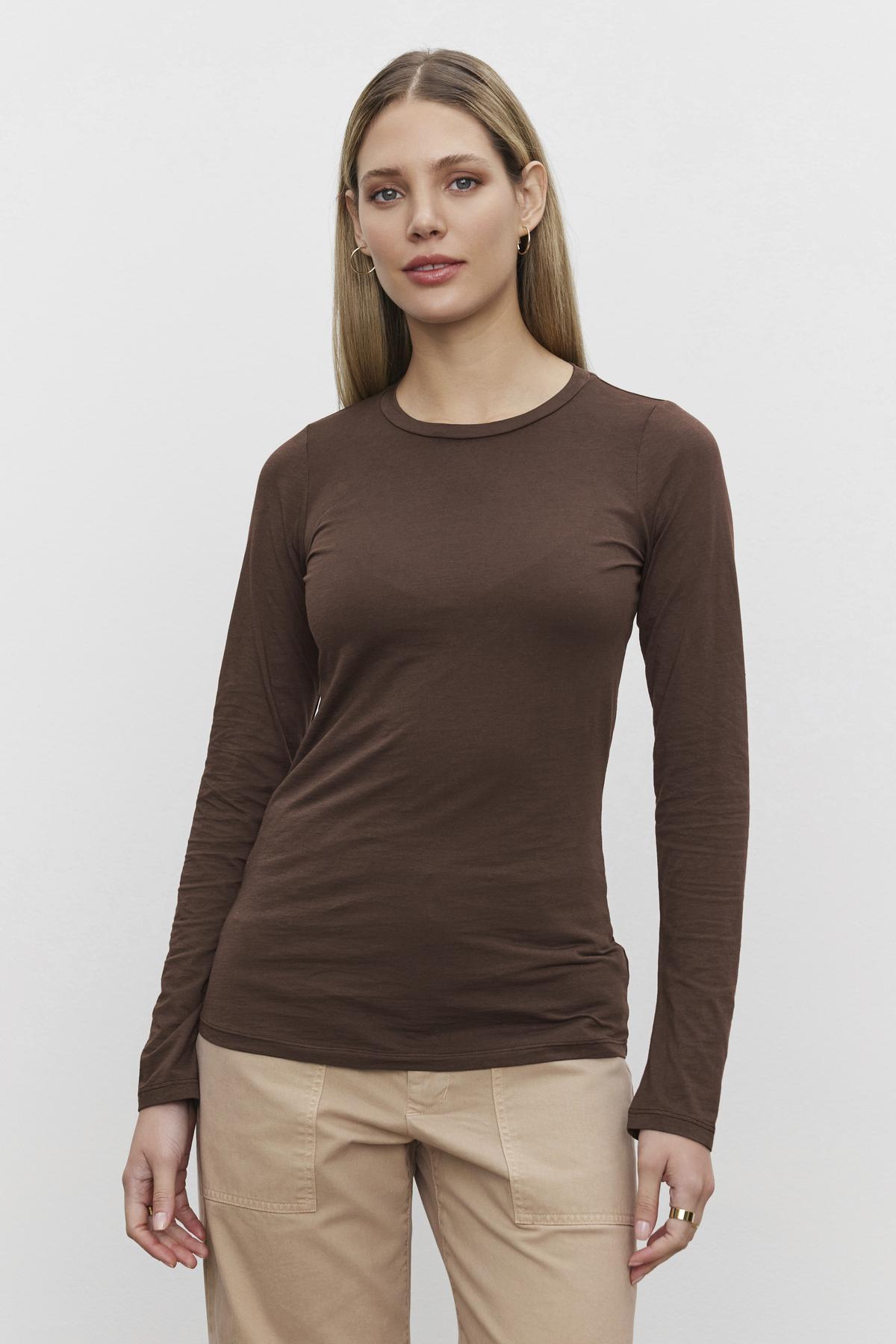   A person with long hair wearing an ultra-soft ZOFINA TEE in whisper brown and beige pants stands against a plain white background. The shirt is from the brand Velvet by Graham & Spencer. 