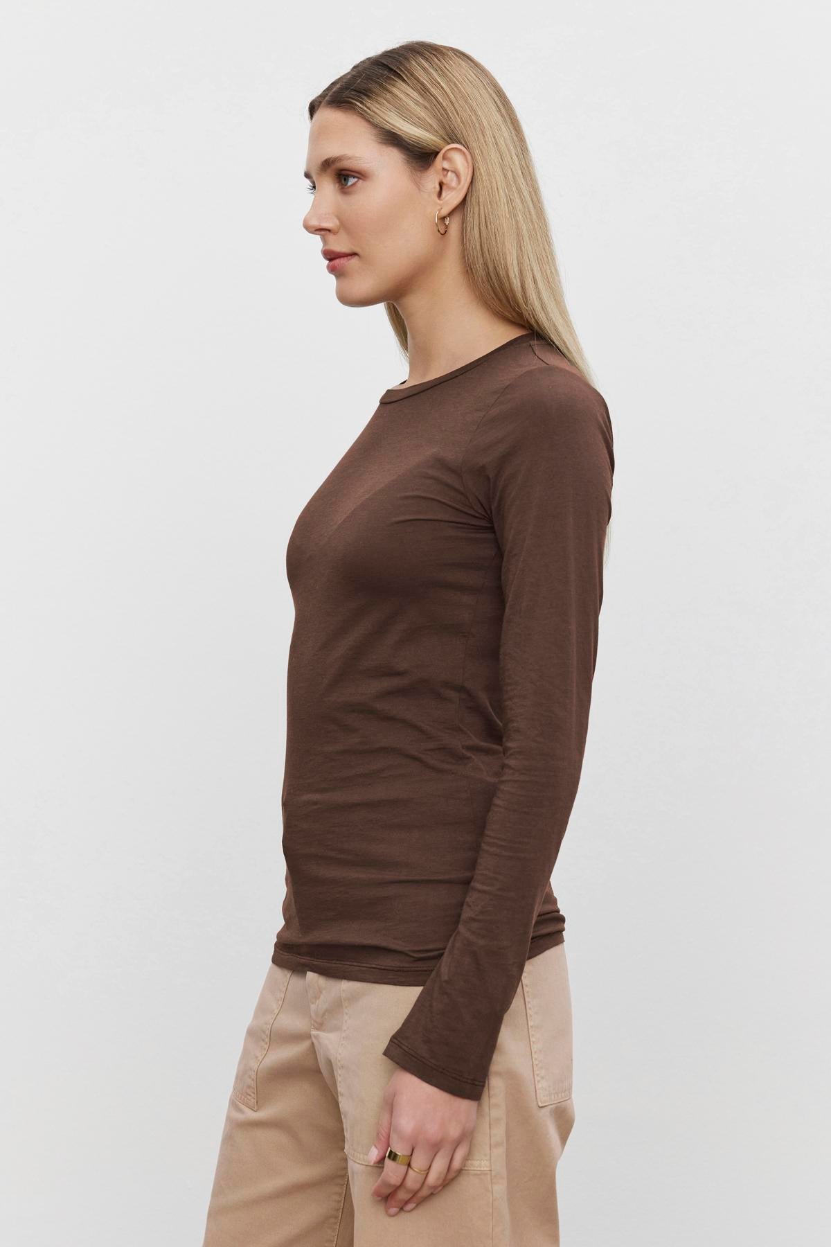 A person with long, light hair is wearing a Velvet by Graham & Spencer ZOFINA TEE and beige pants, standing in profile against a plain white background.-37648688283841