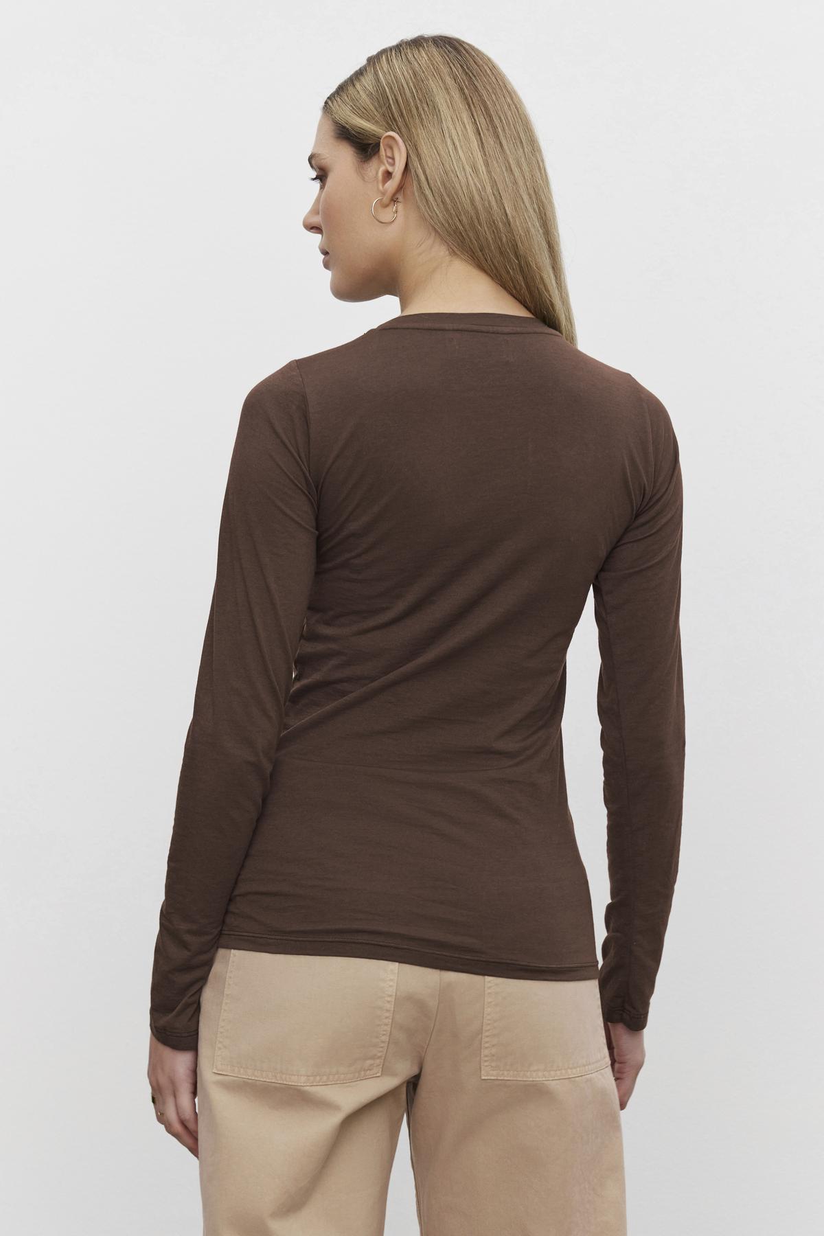   A person with long straight hair is standing and facing away, wearing an ultra-soft ZOFINA TEE by Velvet by Graham & Spencer in whisper brown along with beige pants. 