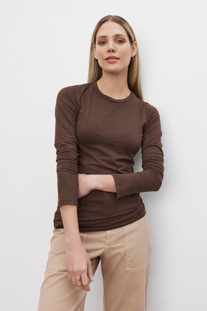 A person with long blonde hair, wearing an ultra-soft ZOFINA TEE by Velvet by Graham & Spencer and beige pants, stands against a plain white background.