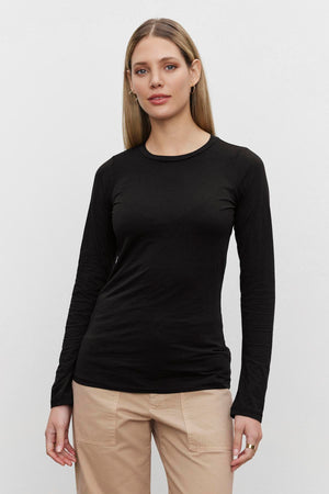 A person with long, straight hair wears the ZOFINA TEE by Velvet by Graham & Spencer, an ultra-soft gauzy whisper fitted black long-sleeve tee with a classic crew neckline, paired with beige pants and stands against a plain white background.