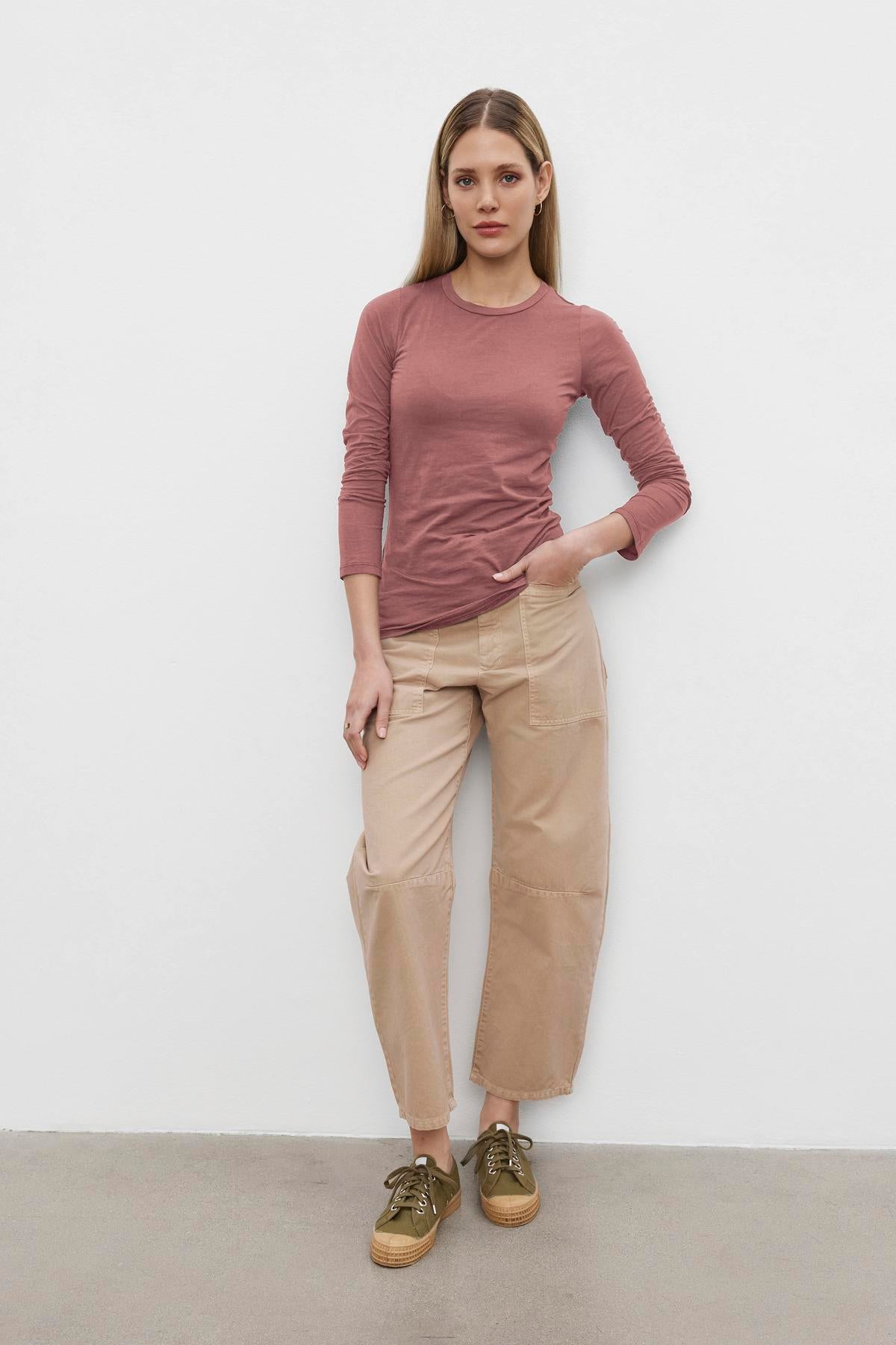   A person with long hair stands against a white wall, wearing the ZOFINA TEE by Velvet by Graham & Spencer in mauve, paired with beige pants and brown sneakers. 