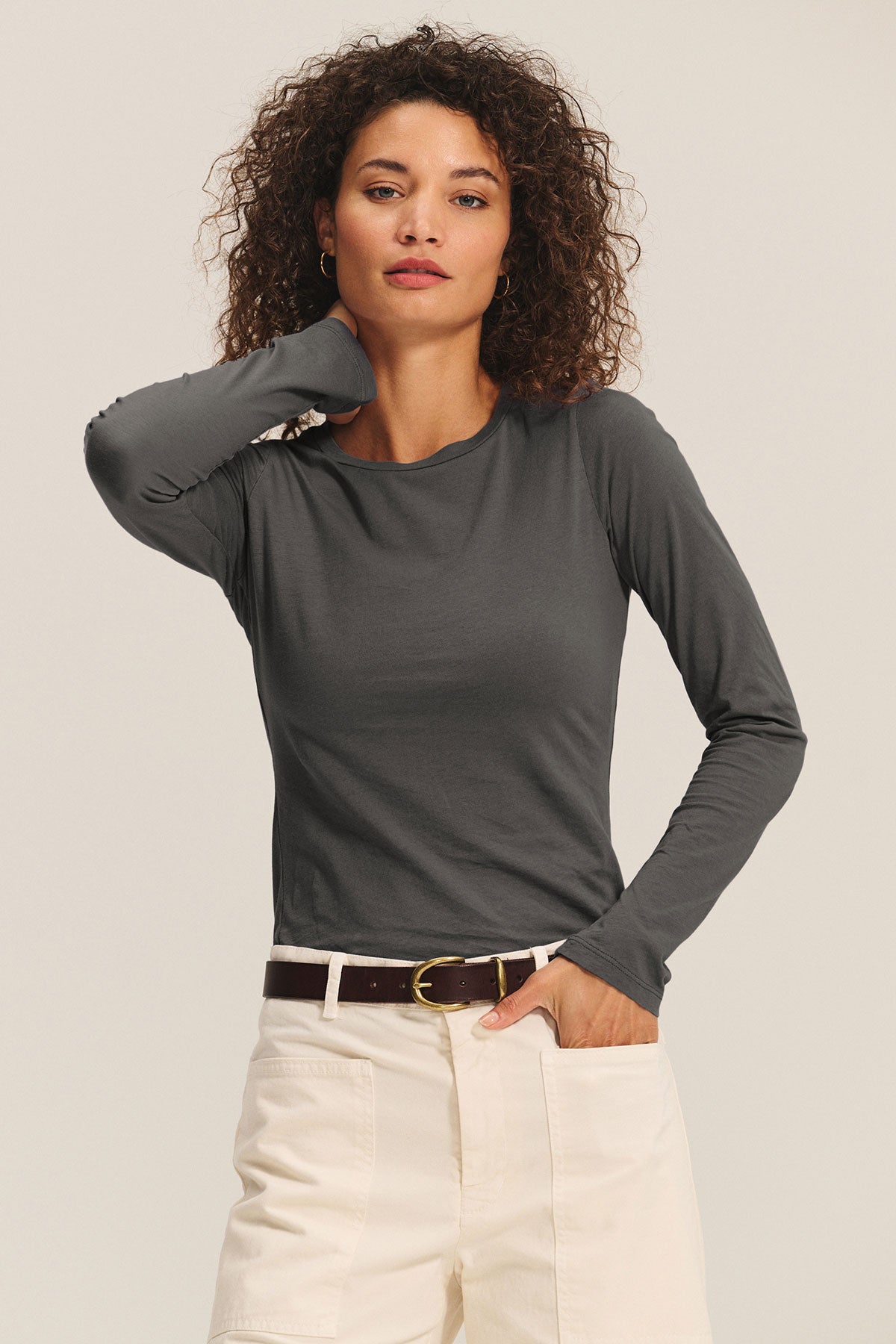 A woman with curly hair wears the ZOFINA TEE by Velvet by Graham & Spencer—an ultra-soft, dark gray fitted long sleeve crewneck—paired with off-white pants, standing with one hand in her pocket against a plain background.-40417102397633