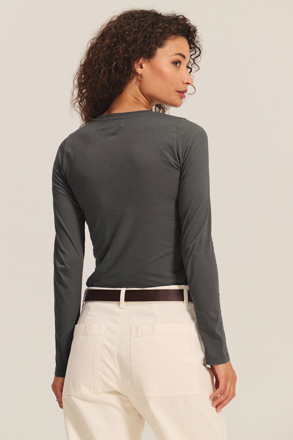 A woman with curly hair is seen from the back against a neutral background wearing the Velvet by Graham & Spencer ZOFINA TEE in dark gray, paired with white pants and a dark belt.